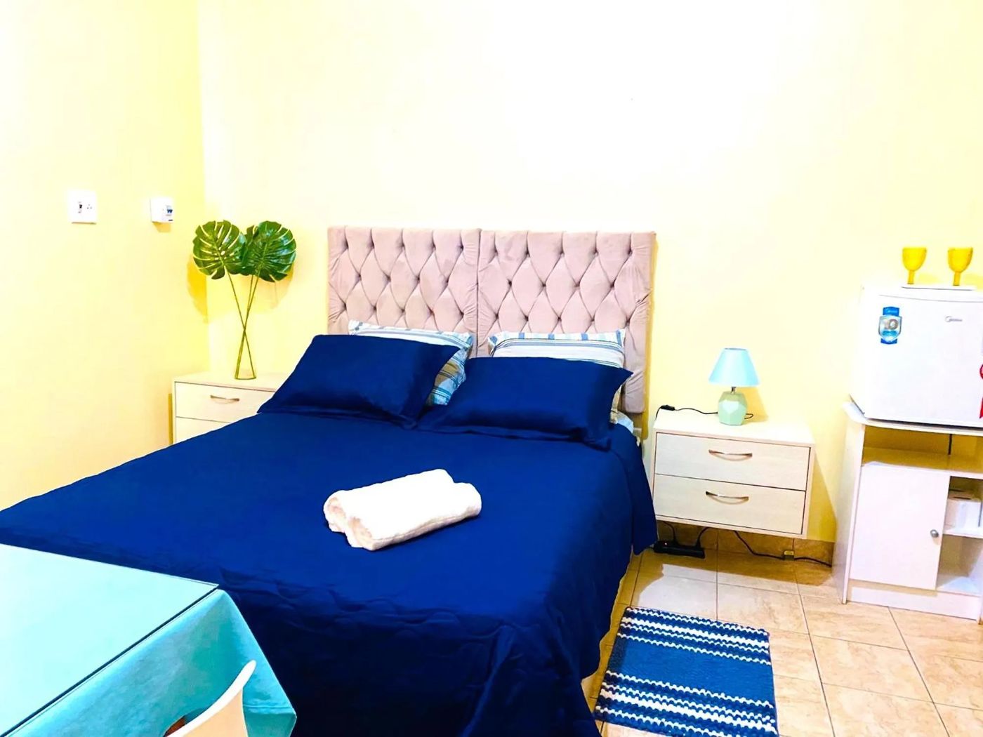 Quarto de casal com roupa de cama azul, frigobar e criado-mudo